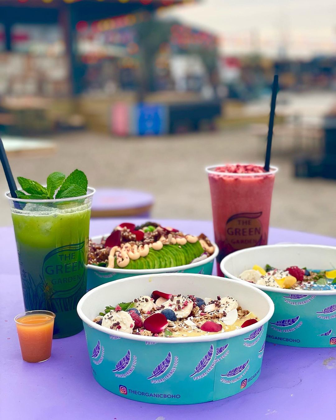 Lebendige, individuell bedruckte Papierschalen, gefüllt mit bunten Smoothie-Bowls, begleitet von erfrischenden Getränken in Plastikbechern mit Logo.